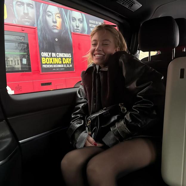 Sydney Sweeney smiling in a car with a red bus advertising a movie in the background.
