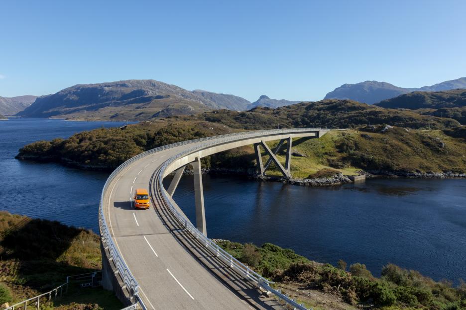 An orange car drives on the Kylesku Bridge over a loch, surrounded by mountains.