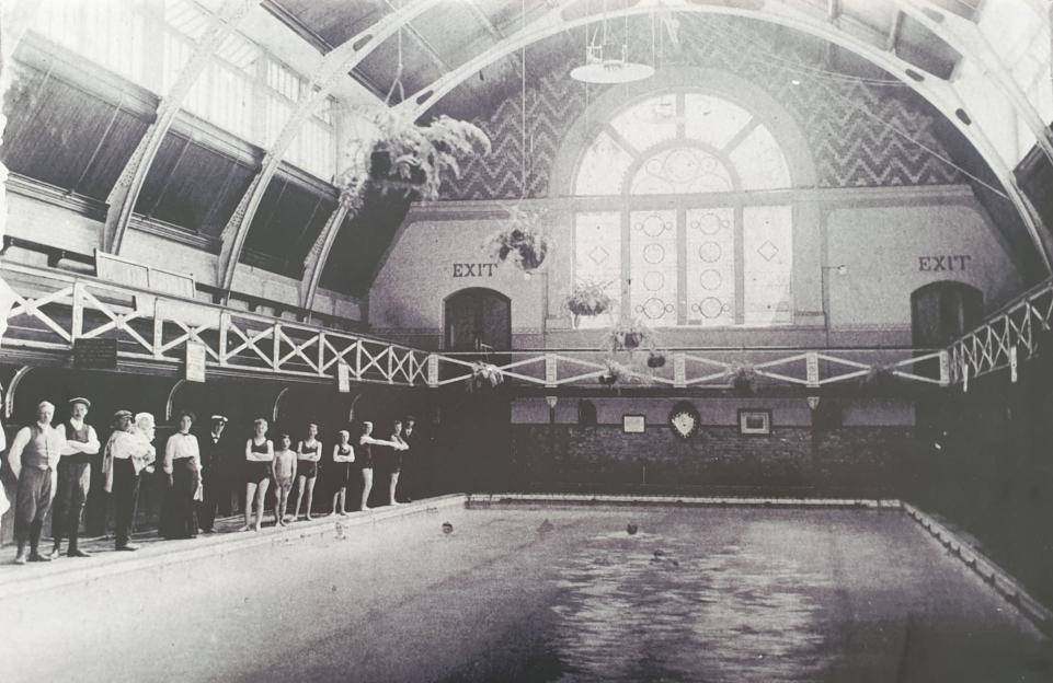 A group of people standing next to a swimming pool in a large indoor facility with ornate architecture.