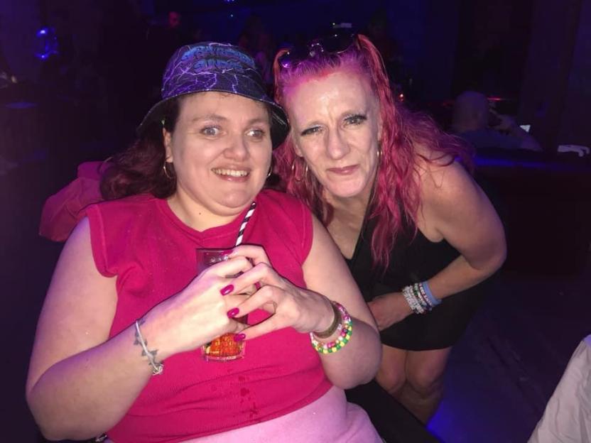 Two women at a club, one with a drink and a patterned bucket hat, the other with pink hair.