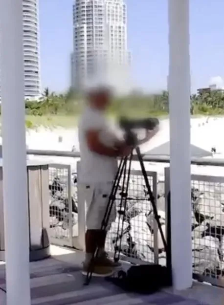 A blurry person standing on a balcony, operating a camera on a tripod, with tall buildings in the background.