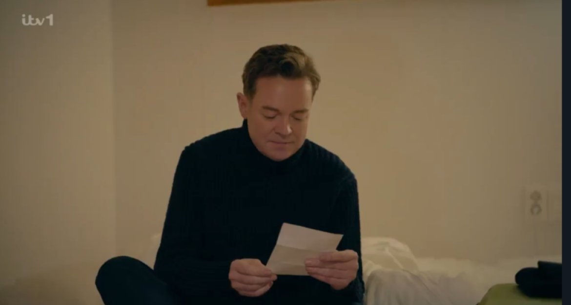 A man in a black turtleneck sitting on a bed reads a letter.