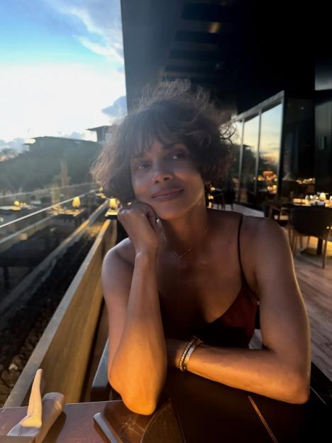 Halle Berry in a red dress with a hand resting on her chin, sitting at a table outdoors at sunset.