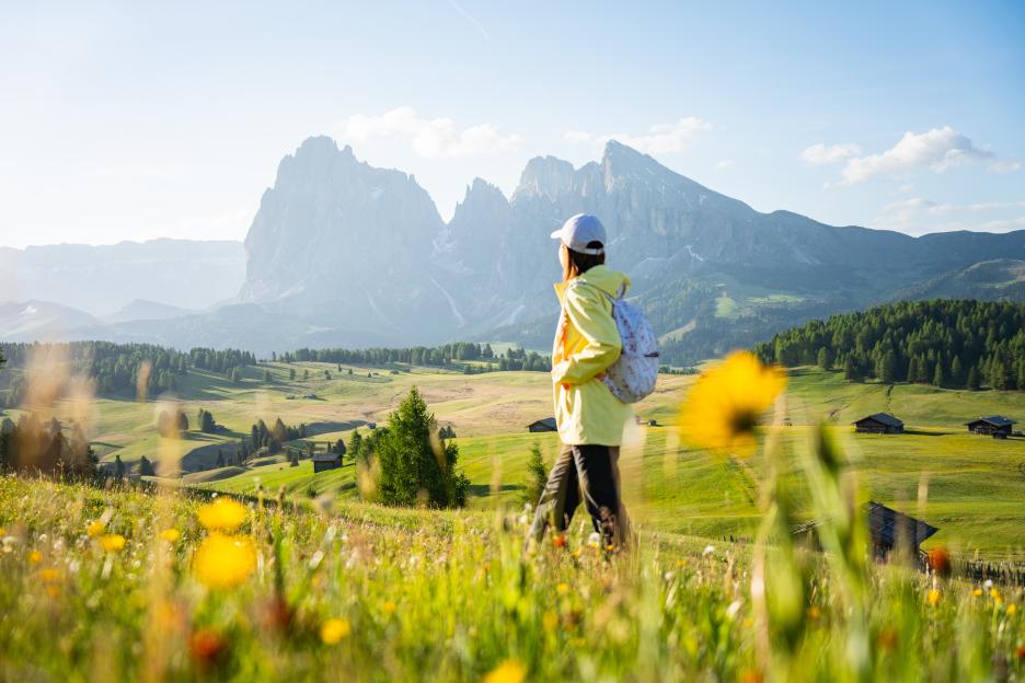 A woman in a white cap and yellow jacket walking through a field with wildflowers and mountains in the background.