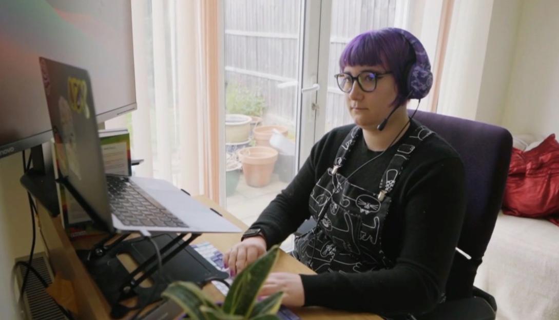 A person with purple hair and glasses wearing a headset and an apron, working on a laptop at a desk.