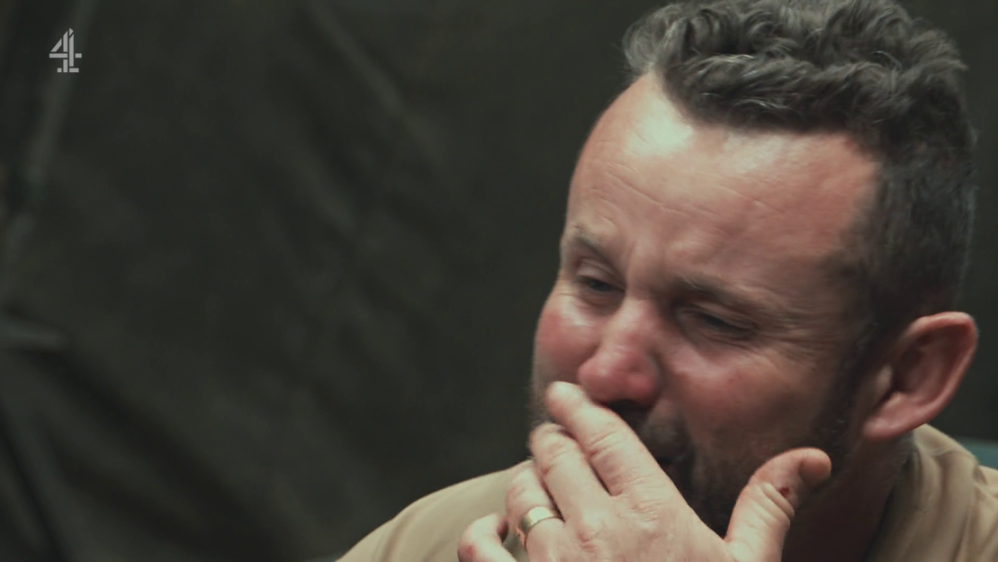 A man with a beard, wearing a ring on his finger, holds his hand to his mouth.