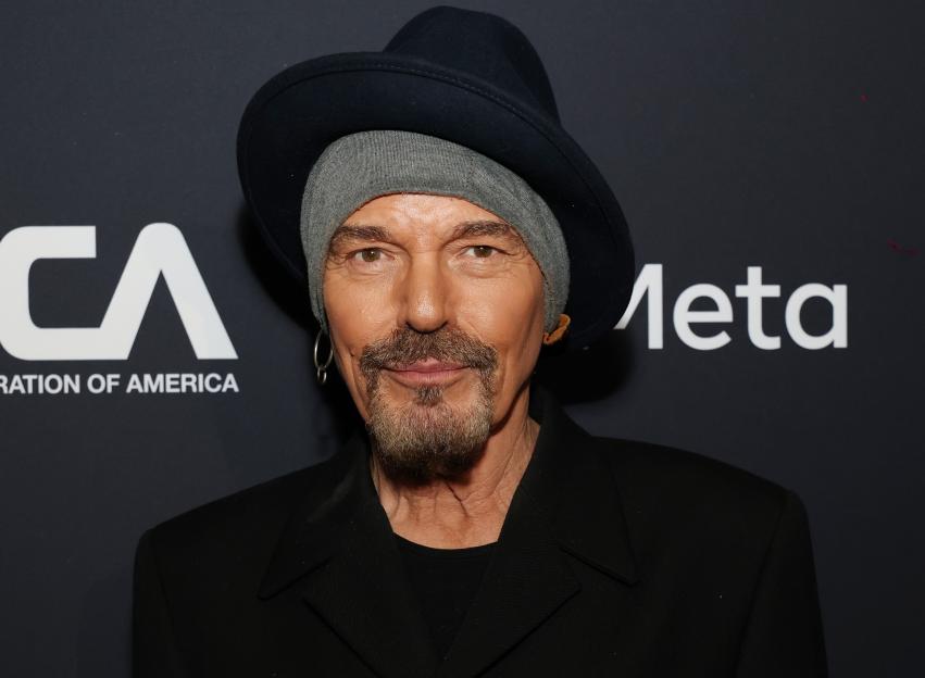 Billy Bob Thornton in a black fedora and grey beanie, black jacket, and goatee.
