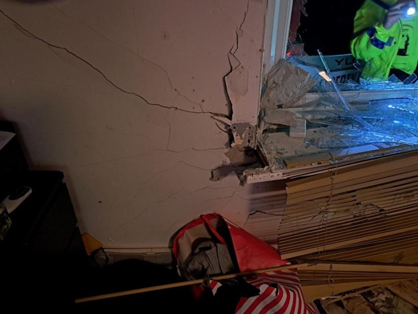 Damaged wall with plaster cracking around a shattered window, showing outside.