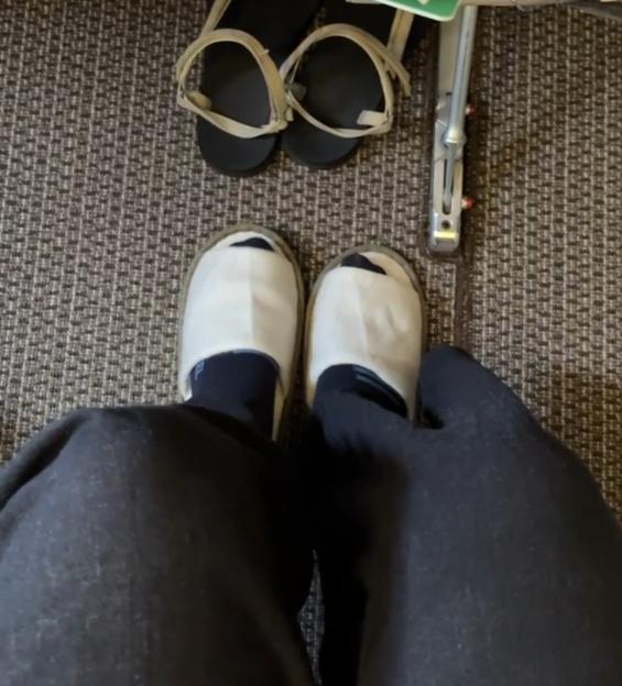 Person's feet in white slippers and dark socks, with their sandals tucked under the seat ahead.