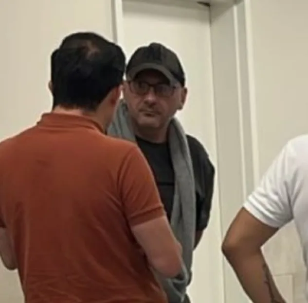 Matthew Ashley Foster-Smith in a black cap and glasses with a grey towel around his neck, looking at a man in a brown shirt.