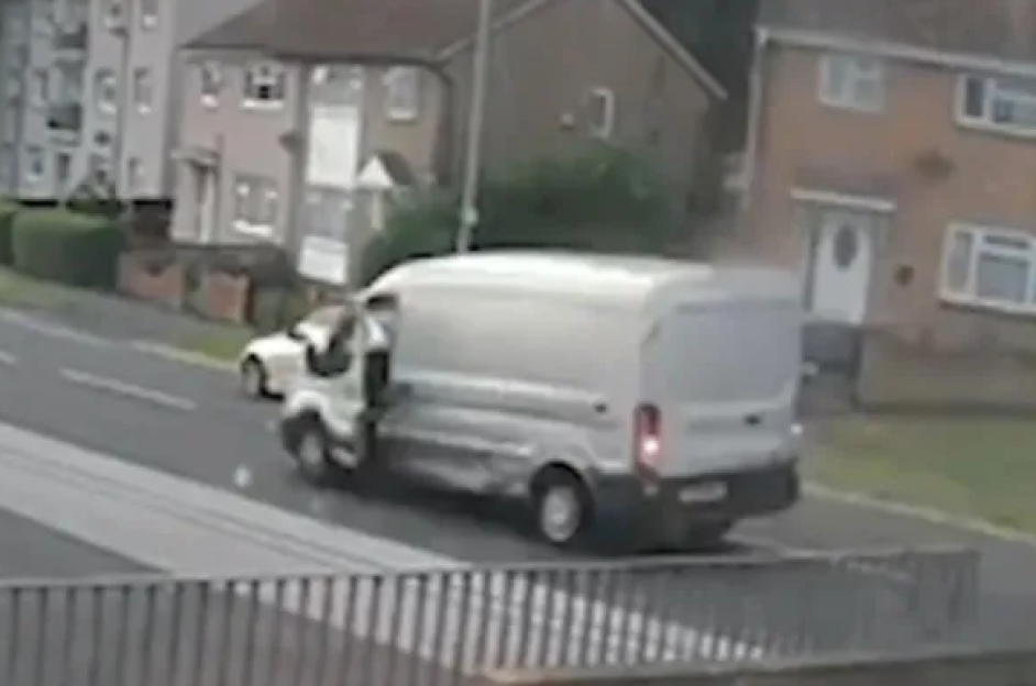 A white Amazon delivery van moving with its side door open, a person hanging onto the side, with residential houses in the background.