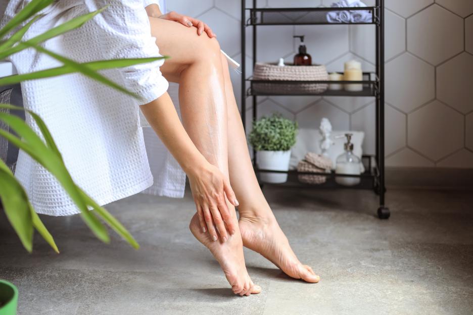 A woman in a white bathrobe applies lotion to her legs in a bathroom.