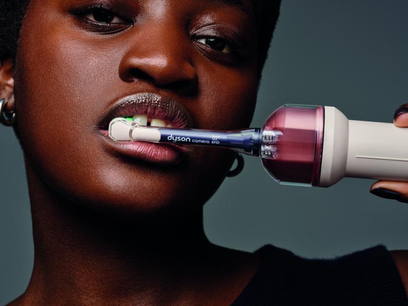A woman with dark skin using a Dyson CameraJet with a green light illuminating her teeth.