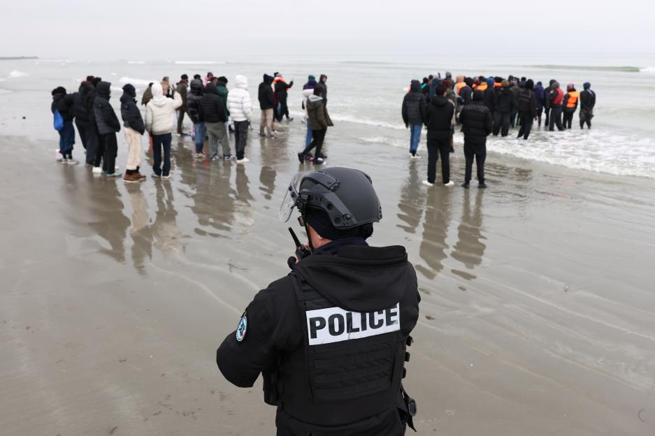 A police officer on a beach watches migrants prepare to board a dinghy to cross the English Channel.