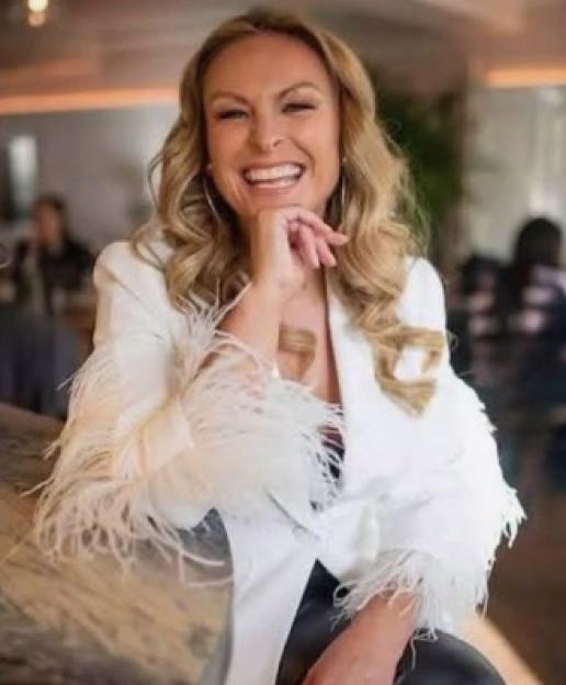 Mel Schilling, a blonde woman, smiling broadly in a white jacket with feather cuffs.