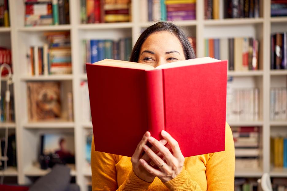 A woman in a yellow sweater smiles while reading a red book in front of a bookshelf.