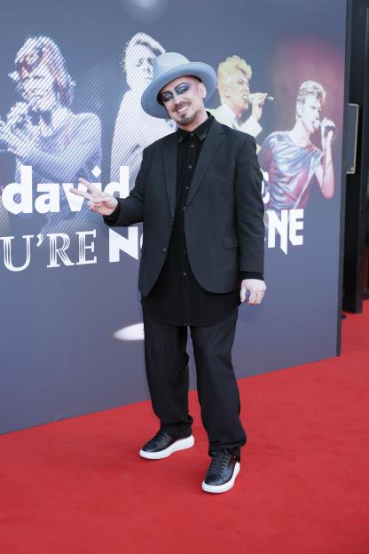 Boy George on a red carpet for the David Bowie: You're Not Alone premiere, making a peace sign.