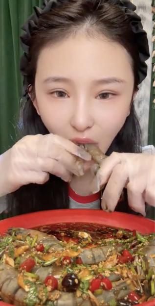 A woman wearing plastic gloves eating a large prawn from a red bowl of seasoned prawns.