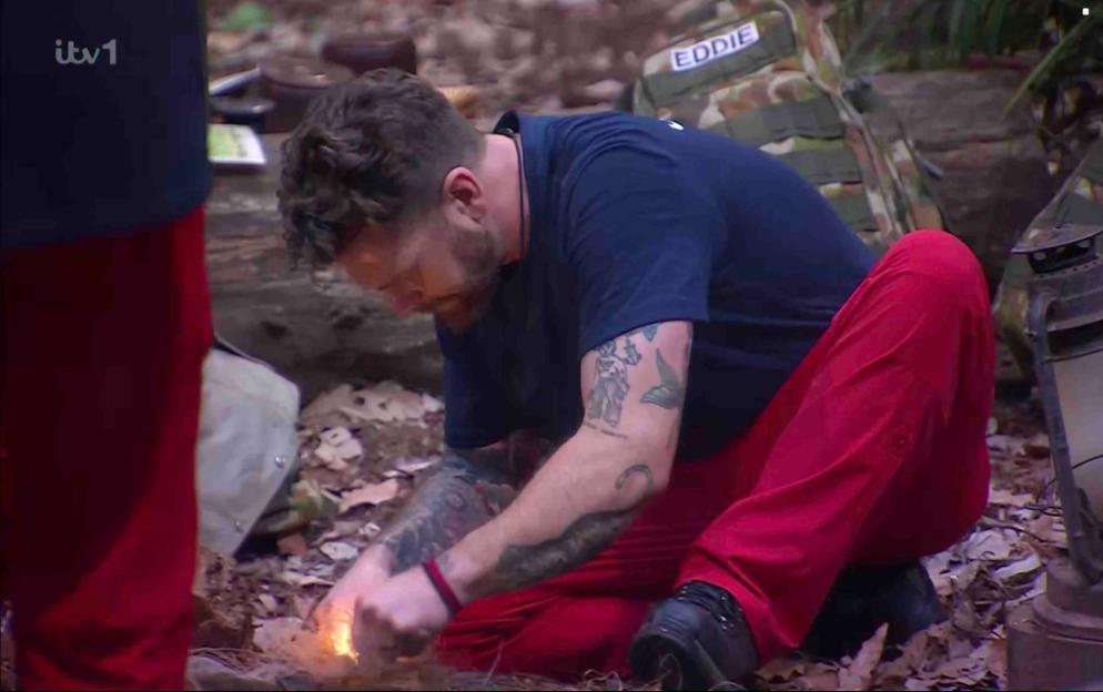Jack Osbourne in a blue t-shirt and red trousers, kneeling and starting a fire.
