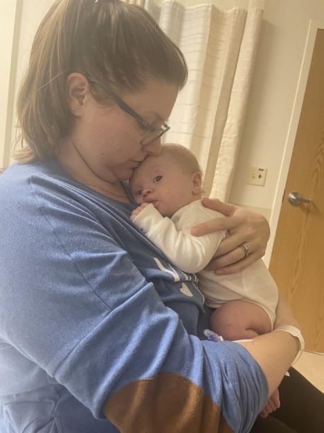 A woman in glasses and a blue shirt holding a baby in a white onesie.