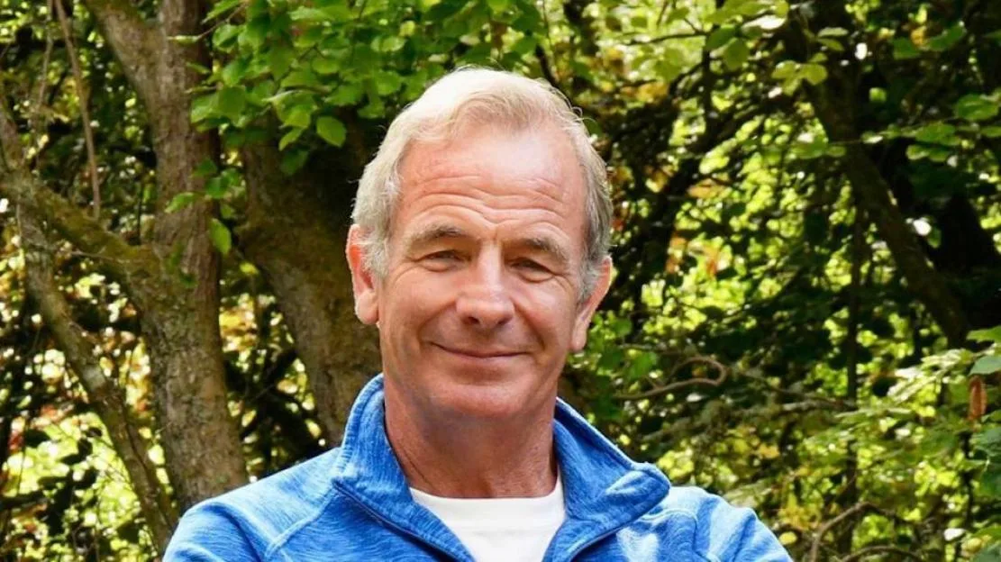 Robson Green standing outdoors in front of a leafy background.