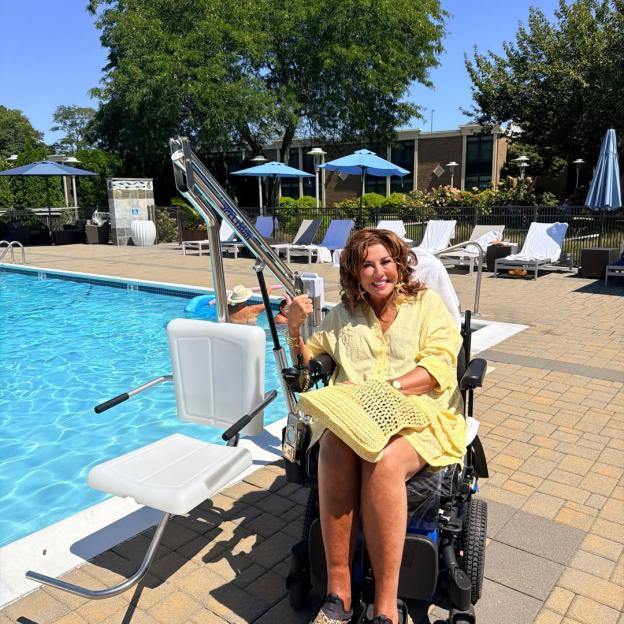 Abby Lee Miller in a yellow swimsuit sitting in her wheelchair next to a pool lift.