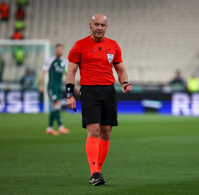 Bald male referee in a red shirt, black shorts, and orange socks on a soccer field.