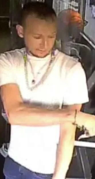 CCTV still of a young man with short hair wearing a white t-shirt and layered necklaces, standing inside a bus.