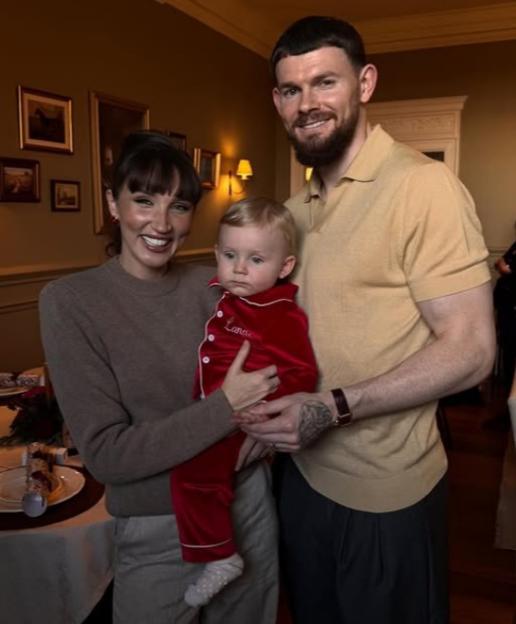A couple and their baby in red pajamas with the name "Lana" embroidered on them, standing in a decorated room.