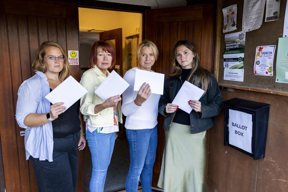 Four women stand outside a ballot box, holding blank pieces of paper representing their votes in a referendum.