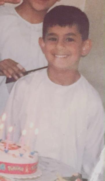 Junaid Ahmed as a child, smiling next to a birthday cake with lit candles.
