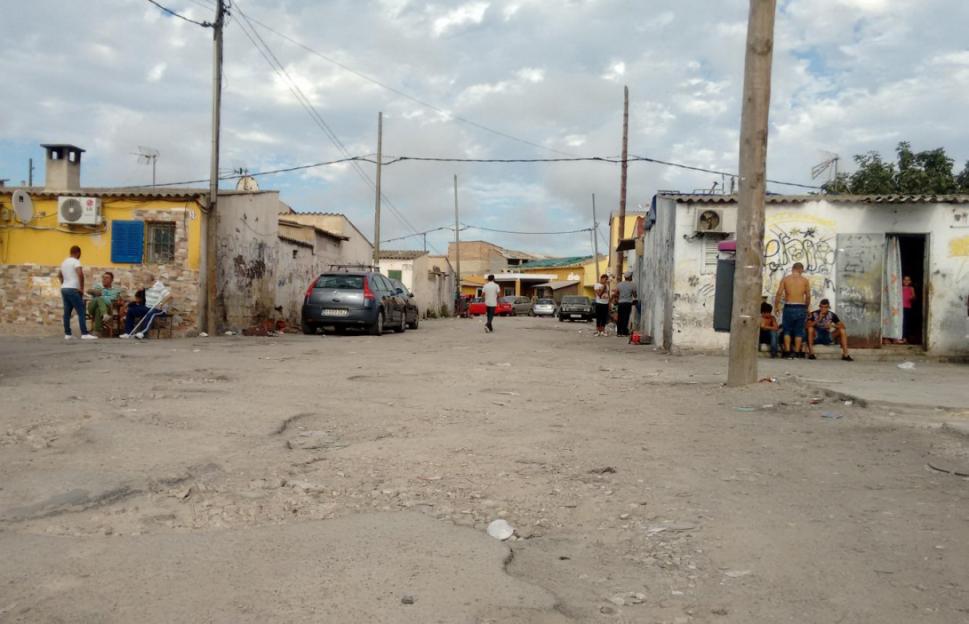 Dirt road in a residential area with people standing and sitting outside houses.
