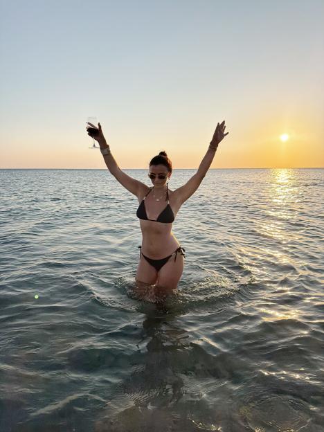Lucy Mecklenburgh standing in the ocean in a bikini with her arms raised, holding a glass of wine, at sunset.