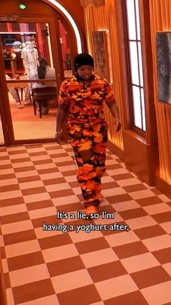 A housemate wearing an orange camouflage outfit walks down a checkered hallway in the Big Brother house.