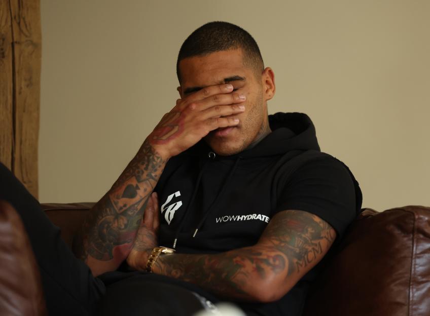 Conor Benn sits on a brown leather sofa, his face covered by his hand, wearing a black hoodie with "WOWHYDRATE" written on it and a gold watch.