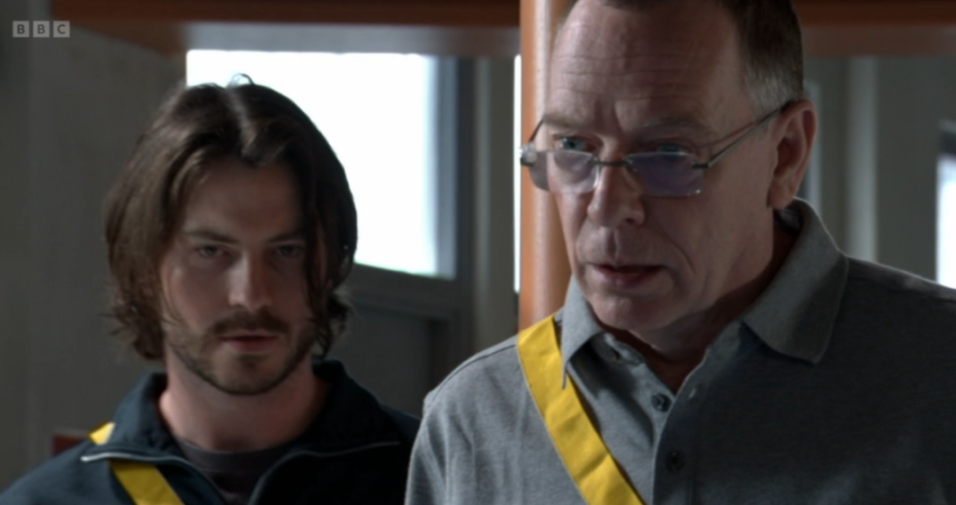 Ian Beale wearing glasses and a gray shirt next to a young man with a beard and dark, long hair.