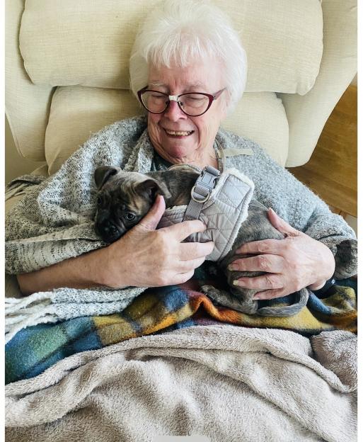 Joyce, a 96-year-old woman, smiles while holding a puppy in a harness.