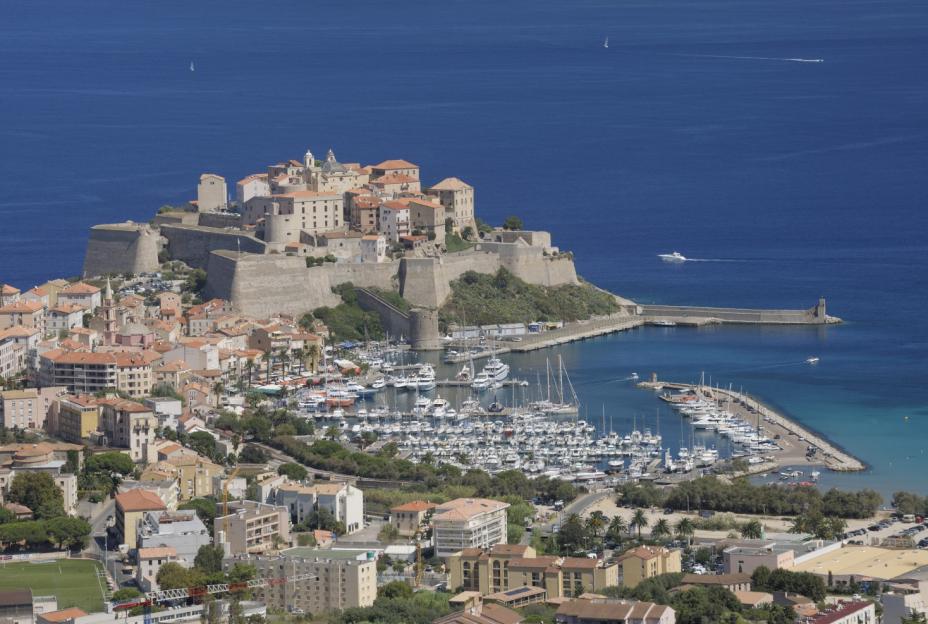 An aerial view of the walled city of Calvi, Corsica, with a harbor full of boats and the Mediterranean Sea in the background.