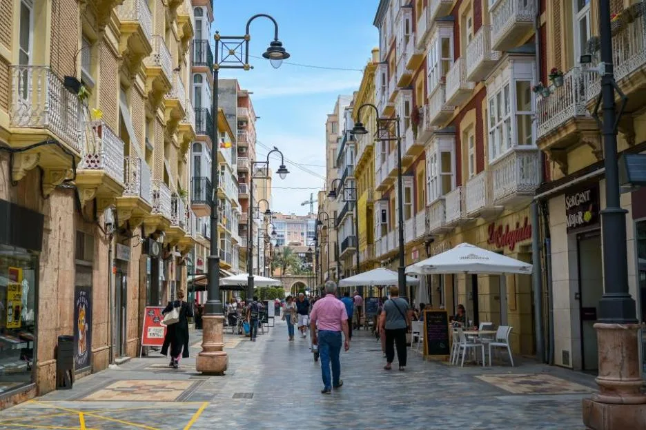 A street scene in Cartagena, Murcia, Spain, showing people walking along a pedestrianized street lined with ornate buildings and outdoor cafes.