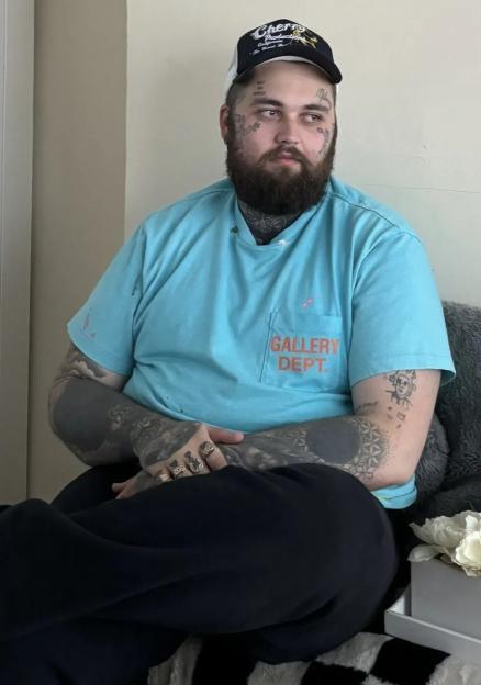 A man with face tattoos, a beard, a blue shirt, and a hat sits on a checkered blanket.