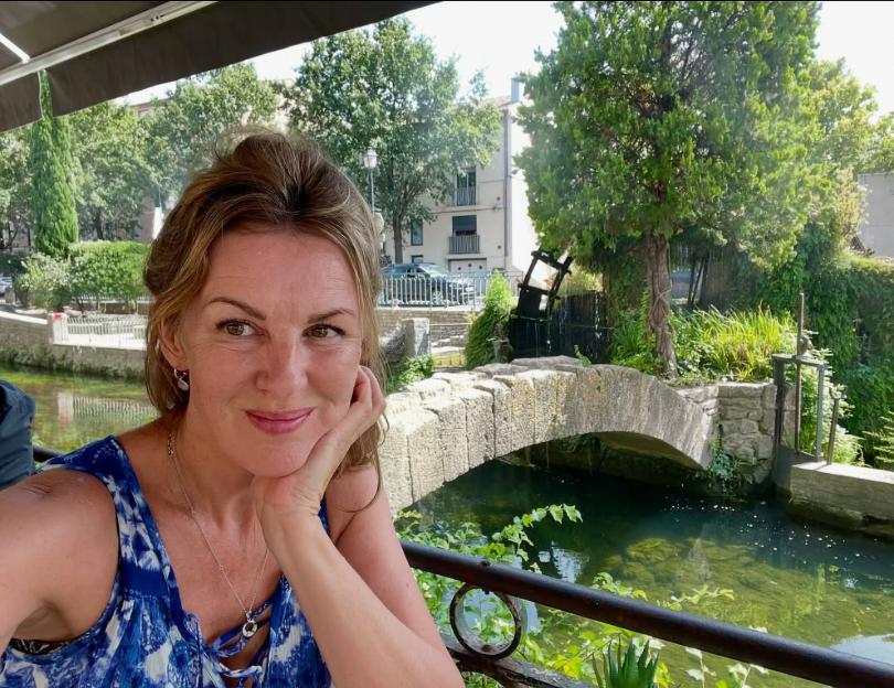 A woman in a blue and white dress rests her chin on her hand, smiling subtly, with a stone bridge over a river in the background.