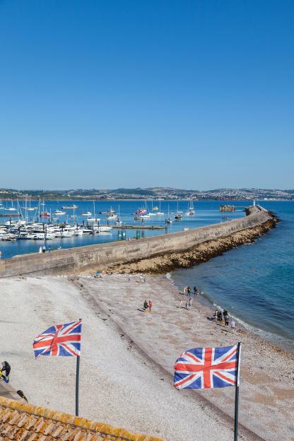 Brixham beach on a sunny summer's morning with boats in the harbor, a long pier, and British flags.