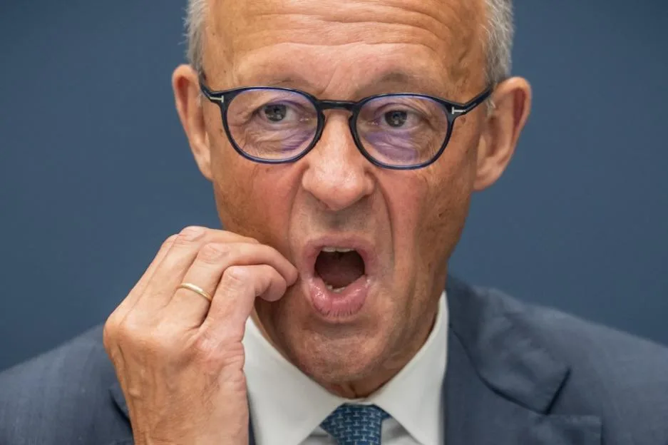 German Chancellor Friedrich Merz, in a suit and glasses, holds his hand to his chin, his mouth open wide.