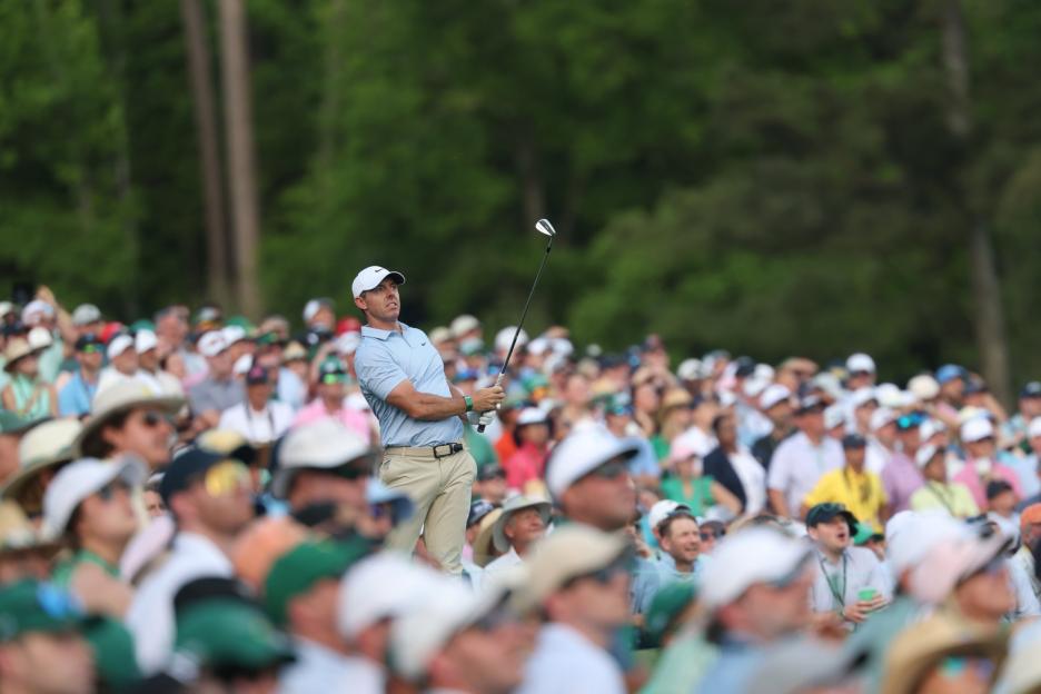 A golfer in a light blue polo shirt and white cap watches his shot with a crowd of spectators in the foreground and background.