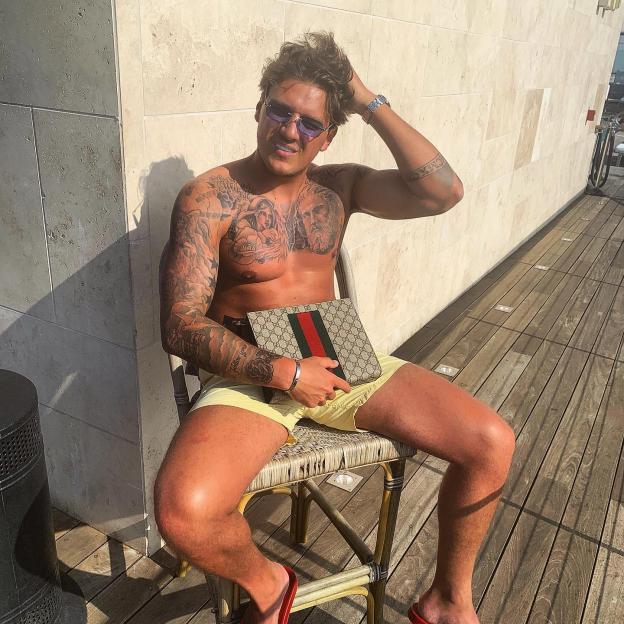 John Joe Slater, shirtless and tattooed, holding a Gucci bag while sitting on a chair.