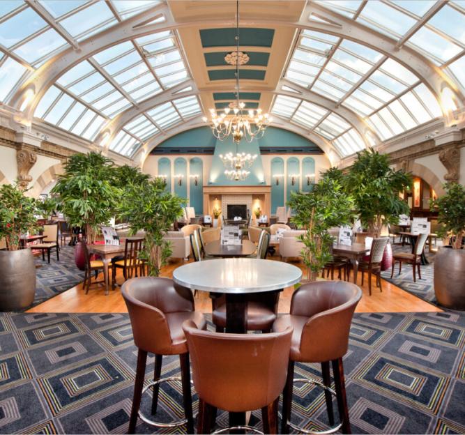 Lobby of a hotel or restaurant with a vaulted glass ceiling, tables, chairs, and potted plants.