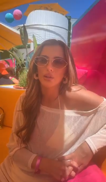 A young woman wearing sunglasses, a white cover-up, and a pink wristband, sitting outdoors.