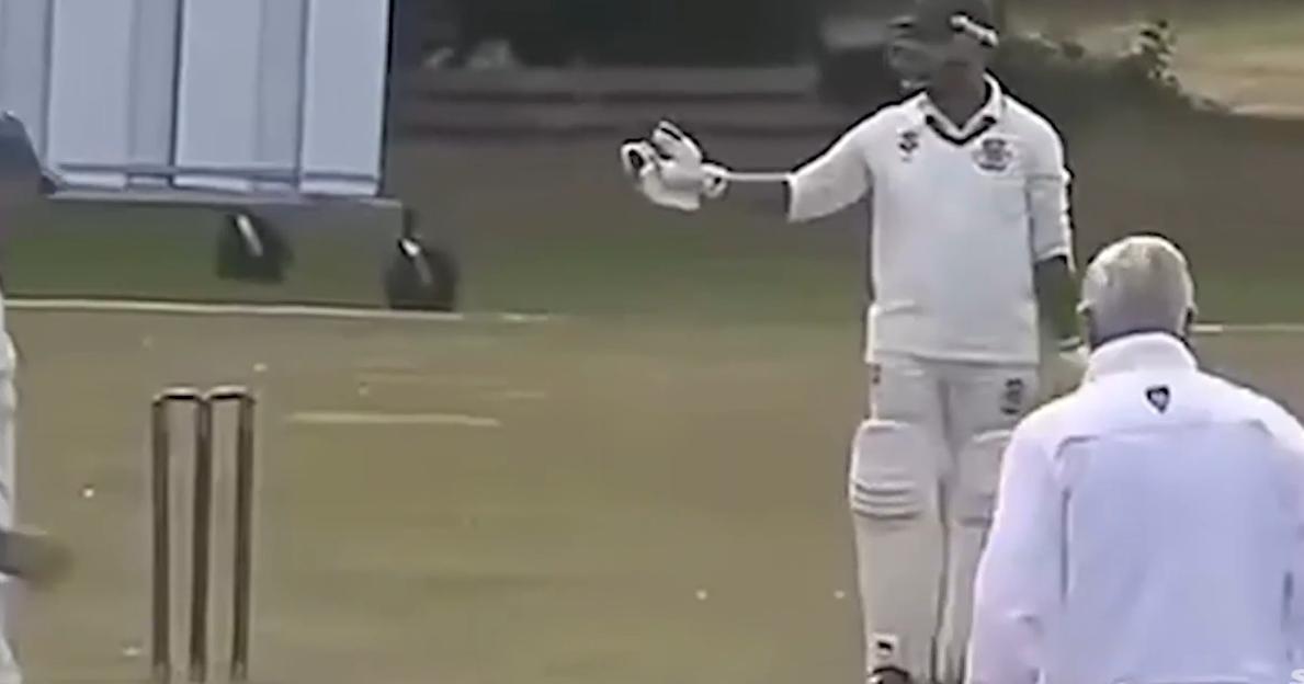 A cricket player in white uniform and helmet holds up a gloved hand while facing an umpire.