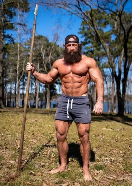 Brian Johnson, known as the Liver King, stands shirtless outdoors holding a spear.