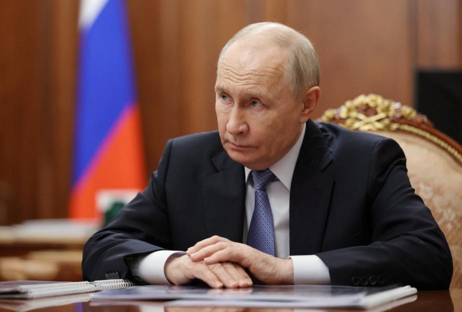 Russia's President Vladimir Putin sitting at a table with hands clasped, looking to the side.